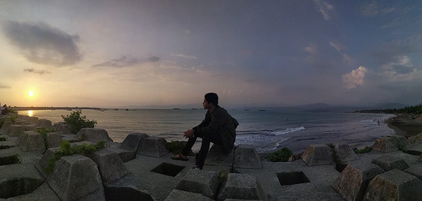 Panoramic sunset view at Batu Bintang with light clouds, featuring Deri Kurniawan sitting on concrete blocks and gazing at the ocean